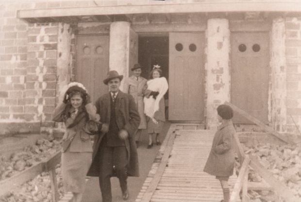 Old black and white photograph, a child and two couples, including a woman holding a baby, are exiting through the front doors of a church under construction.