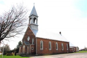 Photographie couleur, plan éloigné, vue latérale d’une église en briques rouges surmontée d’un toit en pente et d’un clocher en tôle. 