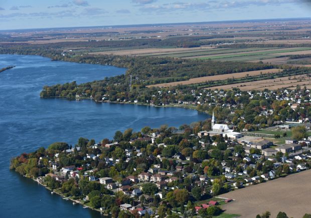 Color photograph, aerial view of an urban area on the waterfront of a large river, in the center, a church and its steeple, in the background, extensive farm lands.