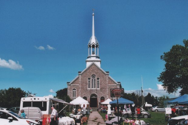 Color photograph, facade of a large stone church in front of which stand people, cars, tables and canopies.