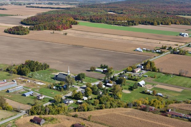 Color photograph, aerial view of a village’s main road, in the center, the church, the cemetery and houses surrounded by farm land.