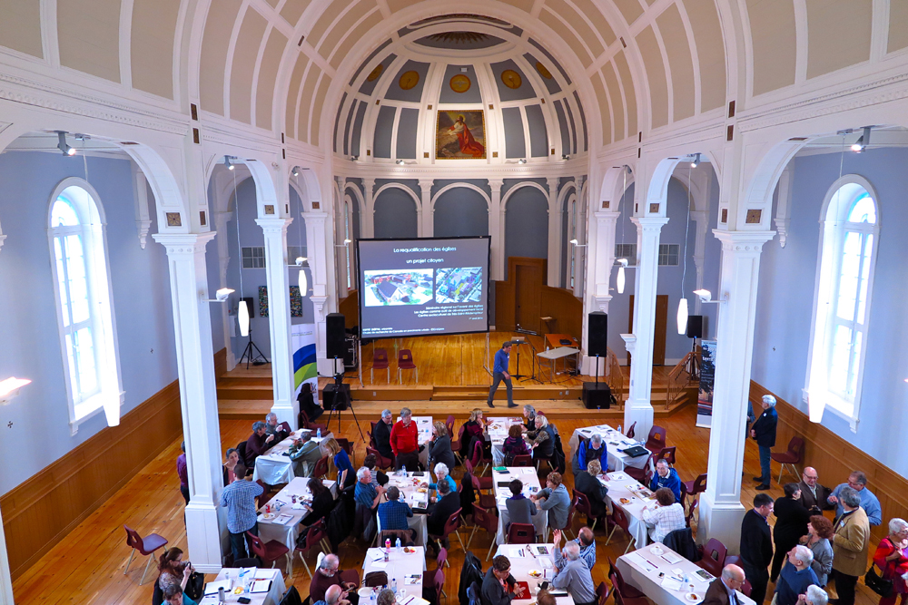 Color photograph, view inside of a church converted into a community center, a vaulted ceiling rests on columns, a man standing in front of a large screen is speaking to a group of people sitting at tables.