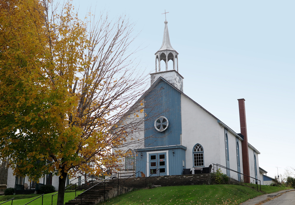 Color photograph, facade of church painted white and blue and whose steeple has no bell, on the front porch, an outdoor furniture set and a dog.