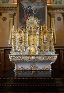 Color photograph, close-up of richly decorated liturgical furnishings which are sculpted and decorated with gold leaf, comprising a tomb and a table on which an upper section holds two statues and wooden gold leaf candlesticks.