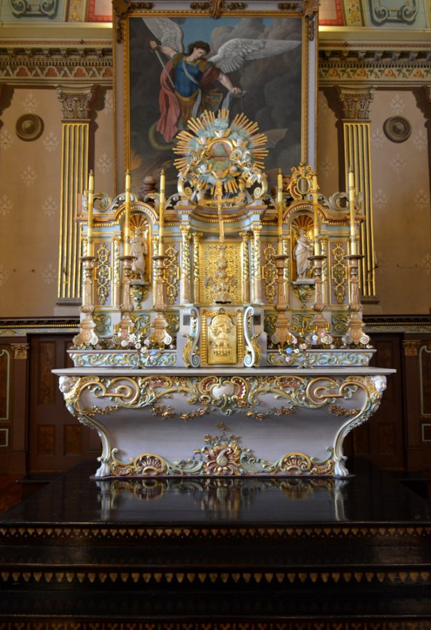 Color photograph, close-up of richly decorated liturgical furnishings which are sculpted and decorated with gold leaf, comprising a tomb and a table on which an upper section holds two statues and wooden gold leaf candlesticks.