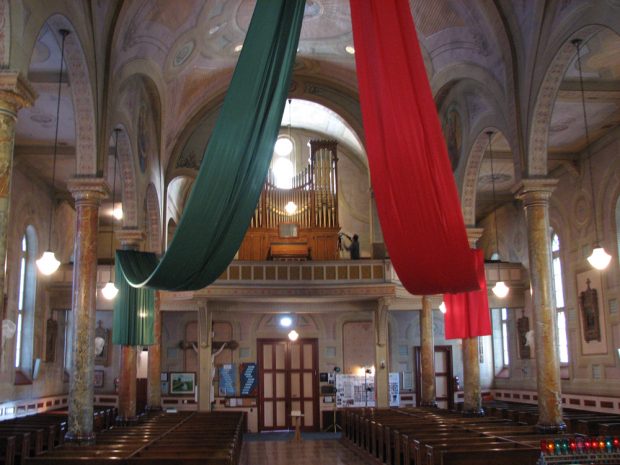 Color photograph, long shot of church interior richly decorated with murals on the walls and columns, two large pieces of fabric green and red are suspended above the nave. On the back gallery, a photograph and a large pipe organ.