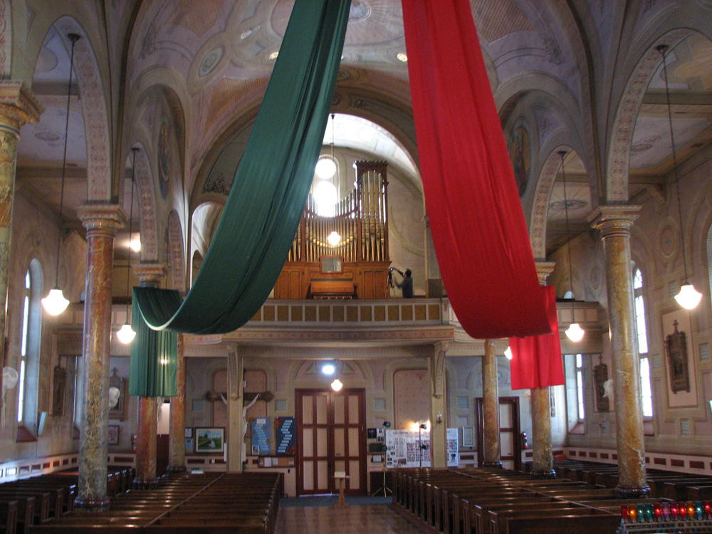 Photographie couleur, plan éloigné, intérieur d’une église richement décorée par des fresques sur les murs et les colonnes, deux grands morceaux de tissus vert et rouge sont suspendus au-dessus de la nef, vue sur la tribune arrière où se trouvent un photographe et un grand orgue à tuyaux.