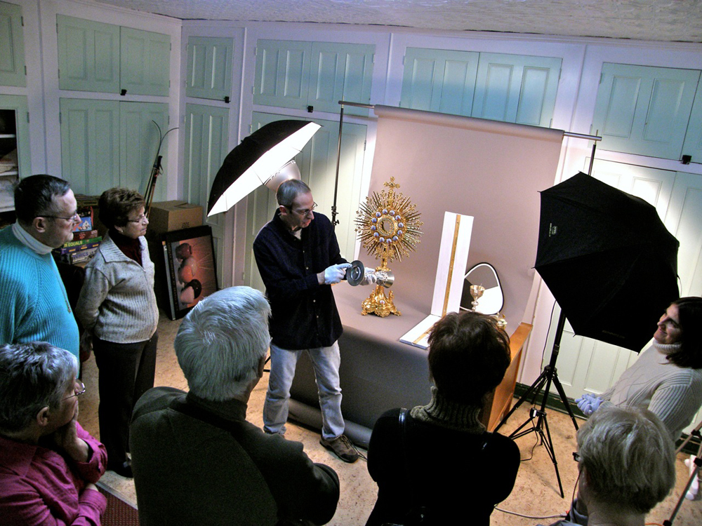 Color photograph, close-up of people listening to a man wearing white gloves and holding an object in his hands, on a table a few religious objects and photographic material.