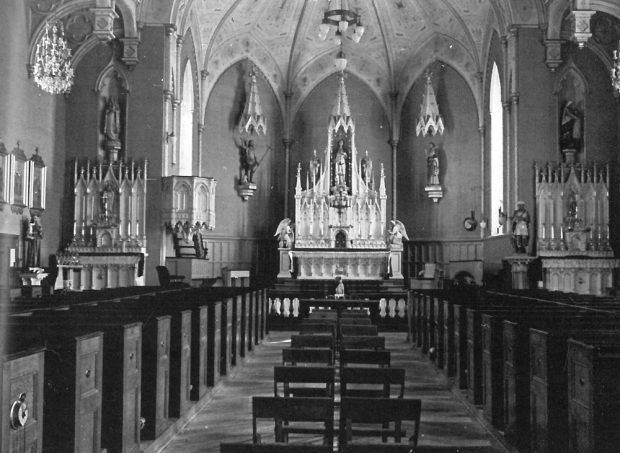 Old black and white photograph, long shot of church interior, in the background, liturgical furnishings and many statues, in the foreground, multiple rows of wooden pews.