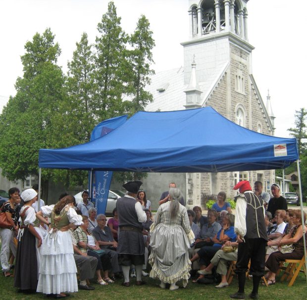 Color photograph, close-up of actors participating in a cultural activity under a canopy between a chapel and a stone church.