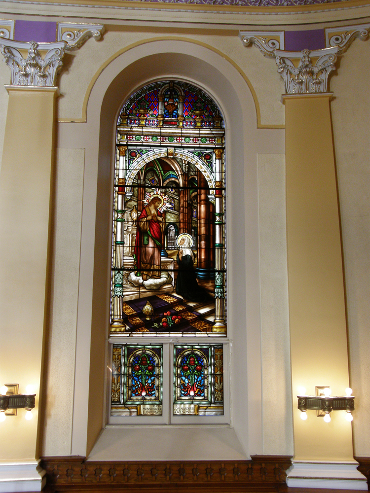 Color photograph, close-up of a long, stained glass oval window showing two characters, a woman kneeling in front of a man standing on a cloud. Near the bottom of the glass an inscription is written: Donated by Mr. Commander Lawrence A. Wilson, 1928, Montreal.