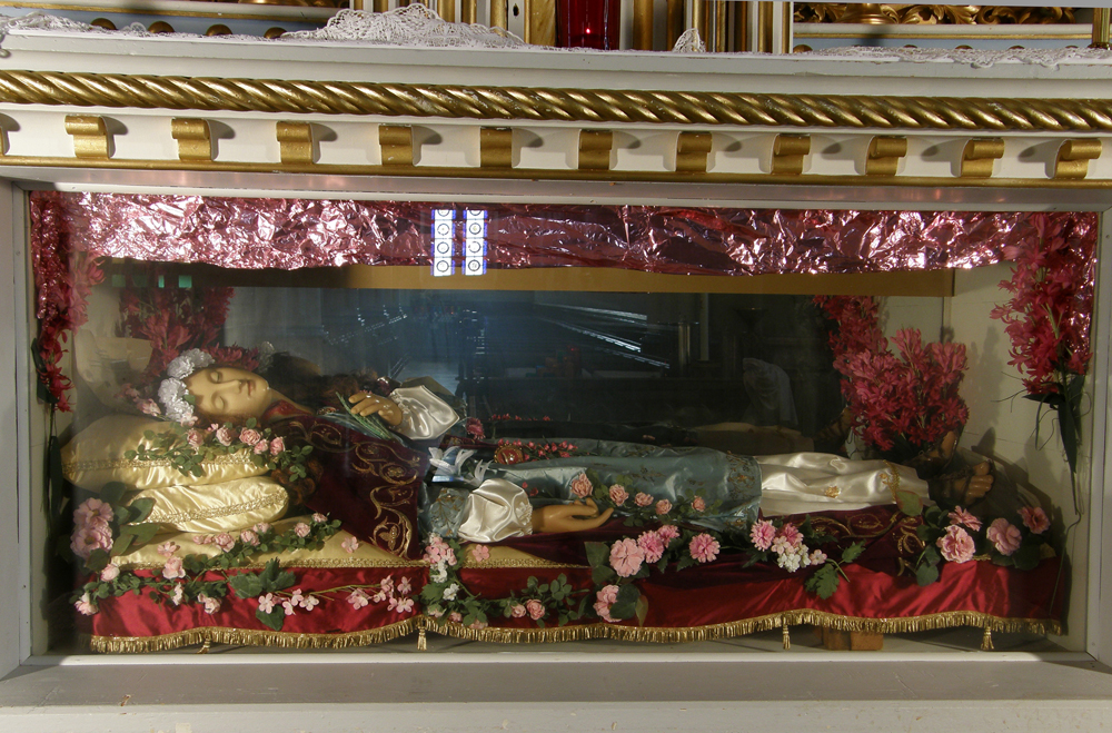 Color photograph, close-up, a life-size wax statue representing a woman dressed in green and white silk is lying on cushions and surrounded by pink flowers in a glass tomb.