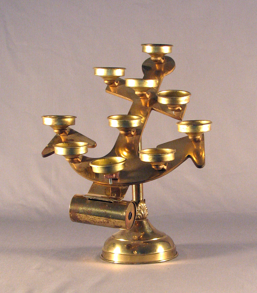 Color photograph, close-up of an anchor-shaped brass religious object with many cells in which can be placed votive candles.