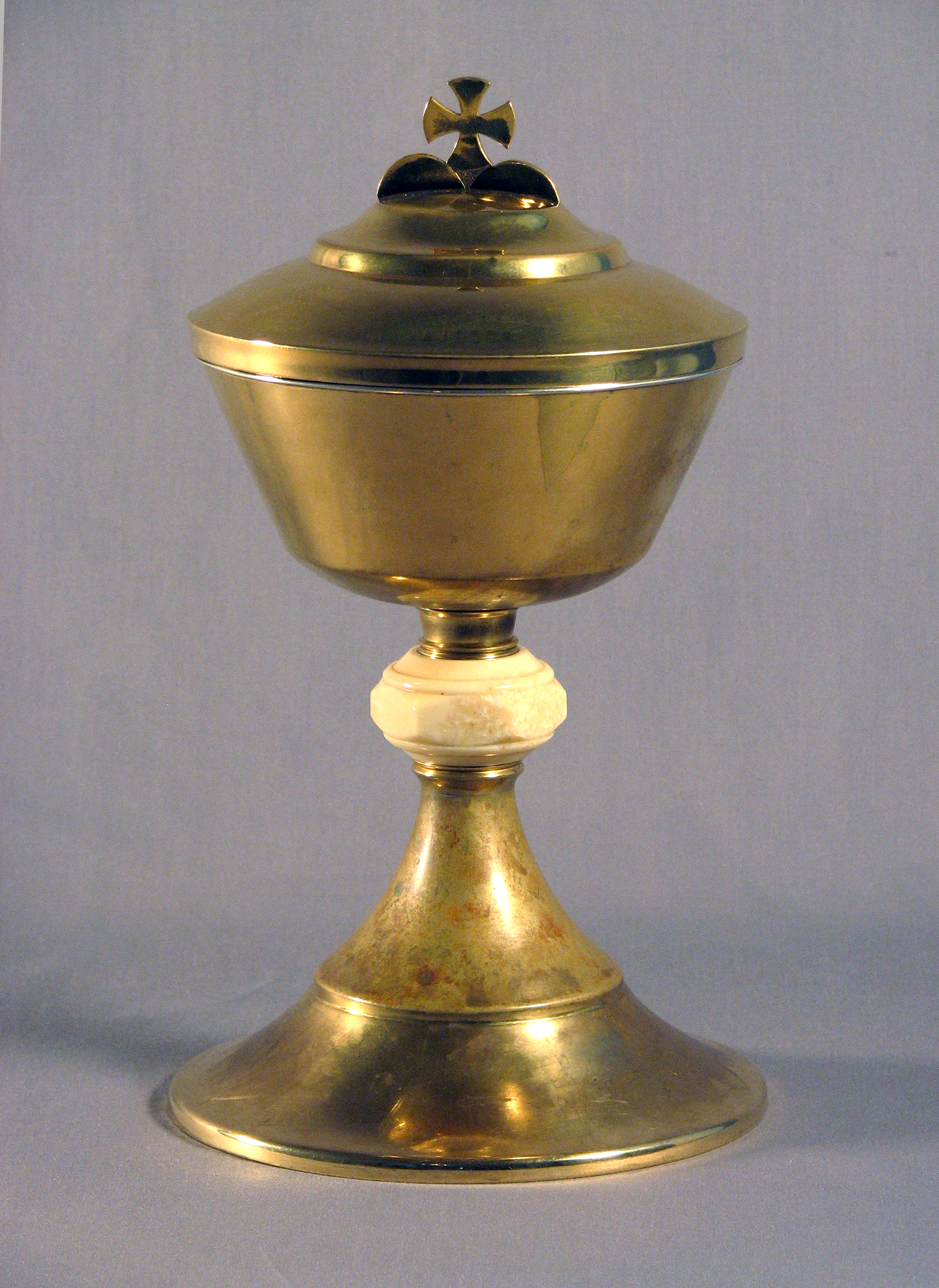 Color photograph, close-up of a religious object made of brass, gold and ivory, in the shape of a stemmed glass and cross-topped lid.