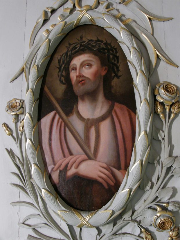 Color photograph, close-up of a painting of a man looking to the heavens and wearing a crown of thorns on his head. The painting is in an oval frame sculpted out of wood and covered in gold leaf.