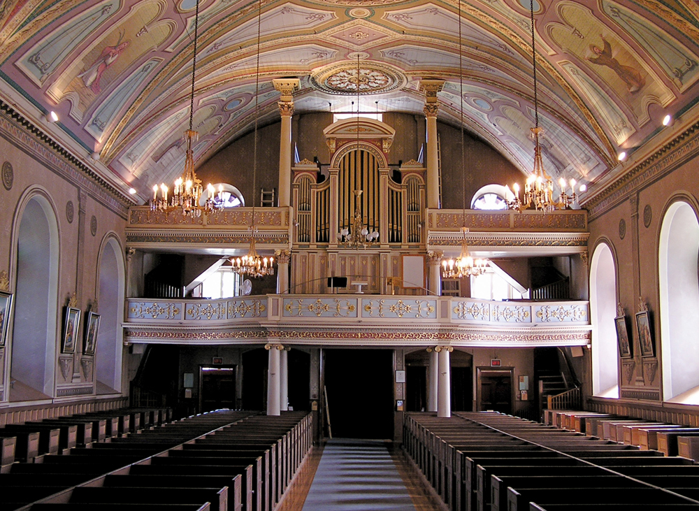 Color photograph, long shot of church interior with decorated dome, in the background a platform and large copper pipe organ, in the foreground, rows of wood pews.