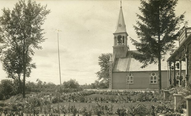 Old black and white photograph, long shot, in the foreground a large flower garden in front of a building, in the background, side-view of a church and its steeple.