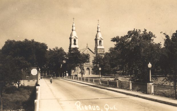 Old black and white photograph, a cyclist on a paved wide bridge leading to a large church with two steeples and surrounded by trees.