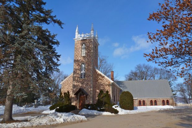 Color photograph, long shot taken in winter, in the foreground, stone church façade with a square steeple and pinnacles, in the background, a river, and a cemetery to the left of the building.