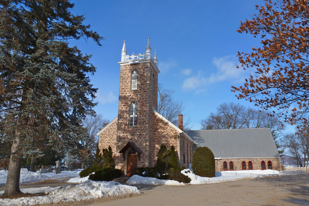 Color photograph, long shot taken in winter, in the foreground, stone church façade with a square steeple and pinnacles, in the background, a river, and a cemetery to the left of the building.