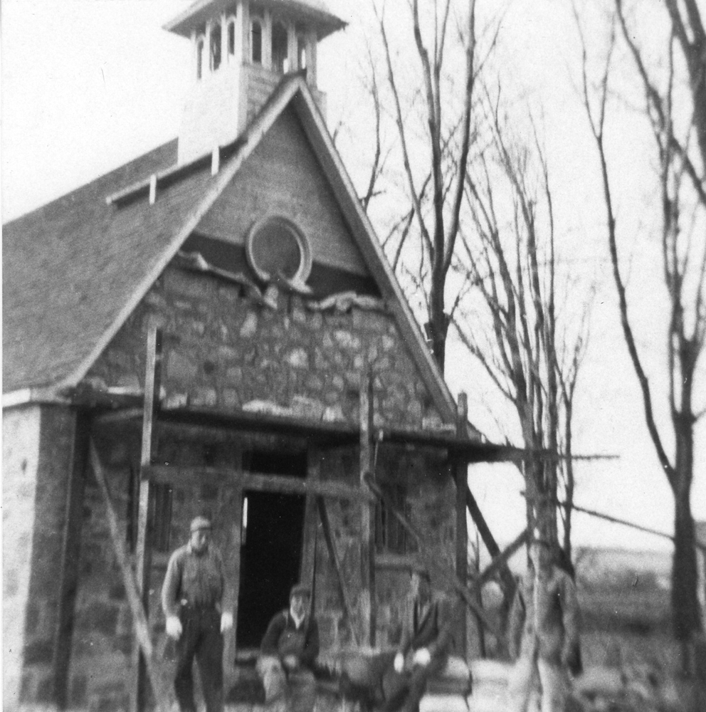 Old black and white photograph, four workers in front of a small stone chapel under construction with scaffolding.