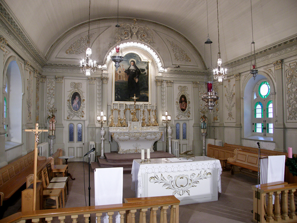 Color photograph, long shot, church interior whose arches and walls are completely covered in white wood panels, in the background, liturgical furnishings and religious artwork, in the foreground, religious furnishings and a varnished wood railing.
