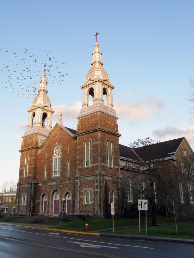 Color photograph, side-view of the façade of an orange brick church topped with two large steeples.