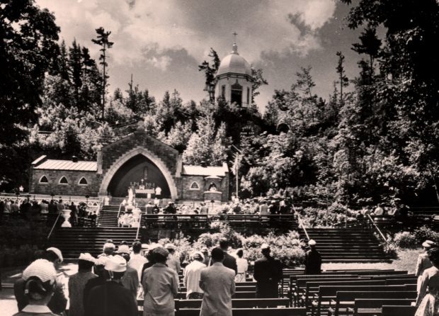 Old black and white photograph, long shot, in the foreground, pilgrims stand at their benches during an outdoor religious ceremony, in the background, an outdoor chapel in the middle of a hill topped with a rotunda and surrounded by trees.