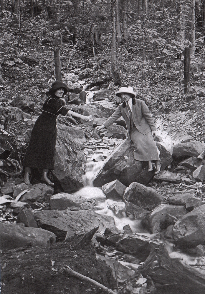 Old black and white photograph, close-up of two women each holding a cup and bending to draw water from a small stream flowing between large boulders.
