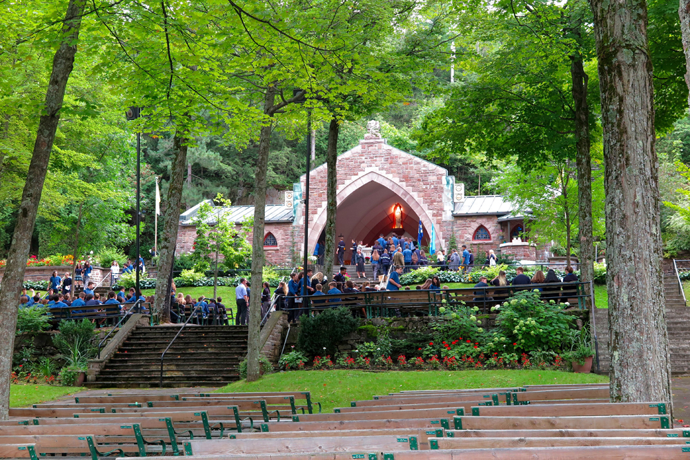 Color photograph, long shot of a crowd assembled outside near an outdoor chapel surrounded by trees and gardens.