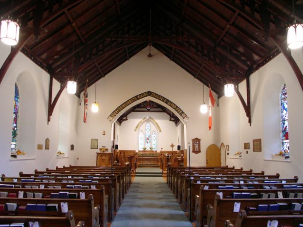 Color photograph, long shot of church interior with a dome entirely sculpted out of wood, in the foreground, wood pews on each side of a main aisle, on the sides, colored stained glass windows, in the background, an atrium with multi-colored windows.