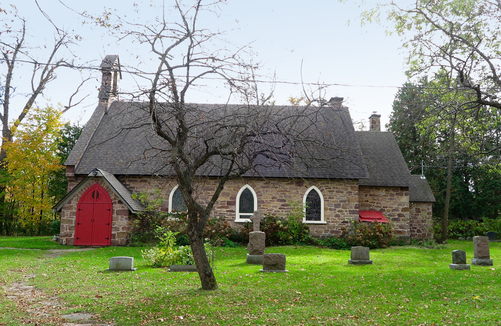 Color photograph, long shot of side-view of a stone church surrounded by trees, with tombstones in the foreground.