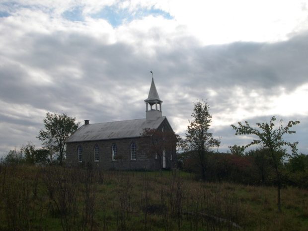 Color photograph, long show of side-view of a small country stone church surrounded by trees and a cloudy sky.