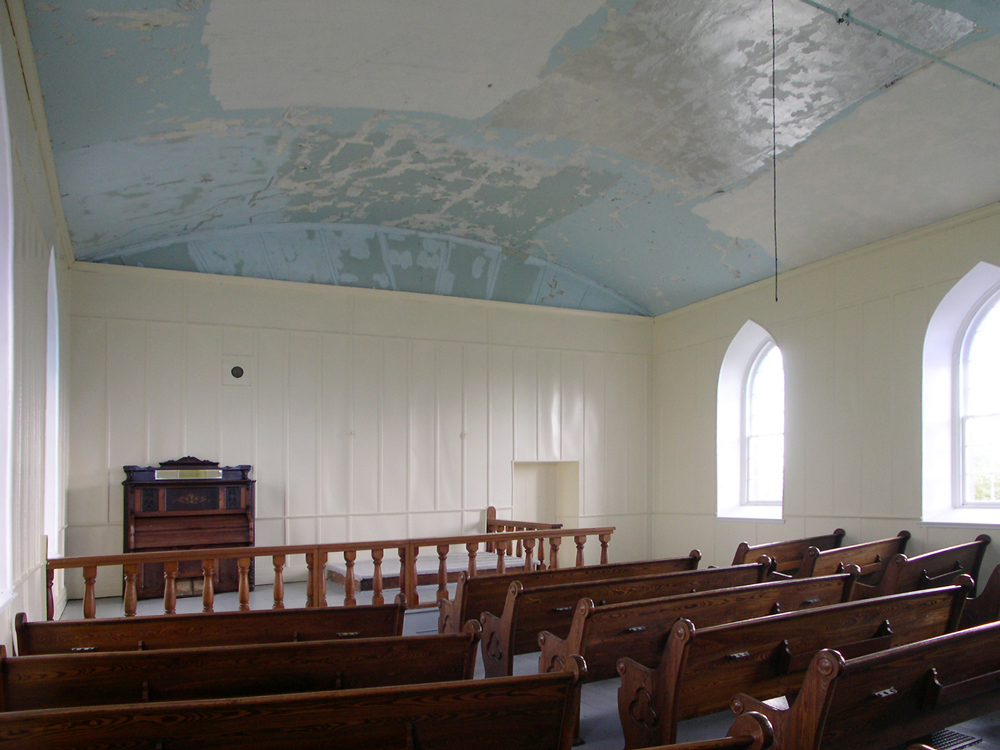 Color photograph, church interior with blank white walls and two windows, in the foreground, wood pews, in the background, a small harmonium surrounded by a wood railing.