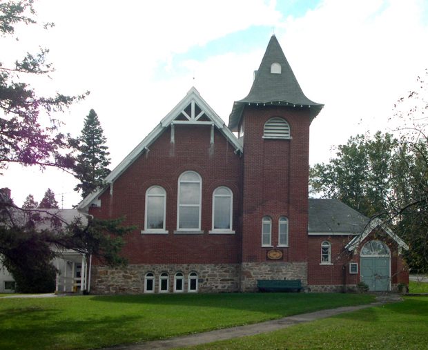 Color photograph, long shot of a red brick church facade with a steep sloped roof and square steeple, in the front, a sidewalk leads to a wood door on the righthand side of the building.