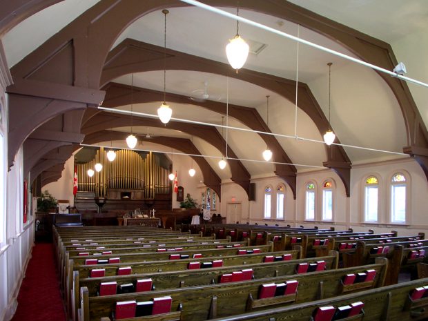 Color photograph, long shot of a church interior with exposed wood beams, in the foreground, wood pews holding red books, in the background, a large copper pipe organ.