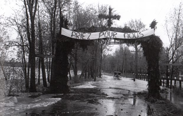 Old black and white photograph, long shot of a car driving on a flooded road bordered by trees above which a cross-topped banner reads: Pray for us.