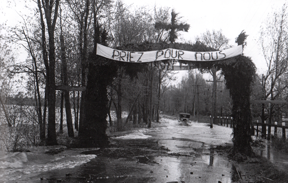 Photographie ancienne en noir et blanc, plan éloigné, une automobile circule sur une route inondée bordée d’arbres au-dessus de laquelle se trouve une bannière surmontée d’une croix sur laquelle on peut lire : priez pour nous. 