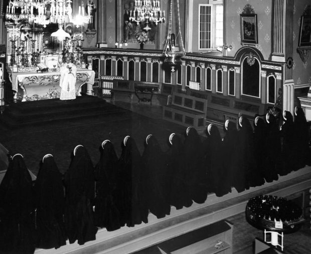 Old black and white photograph, long shot of a richly decorated church interior, in the foreground, nuns all dressed in black are kneeling in front of a celebrant during a religious ceremony.