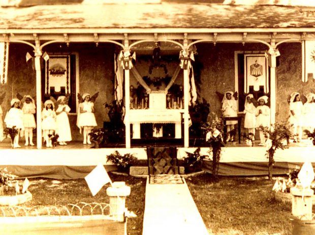 Old sepia photograph, young girls, hands joined, all dressed in white stand on a house porch decorated with flags and religious accessories, the girls personify angels as they stand on each side of an altar of repose.