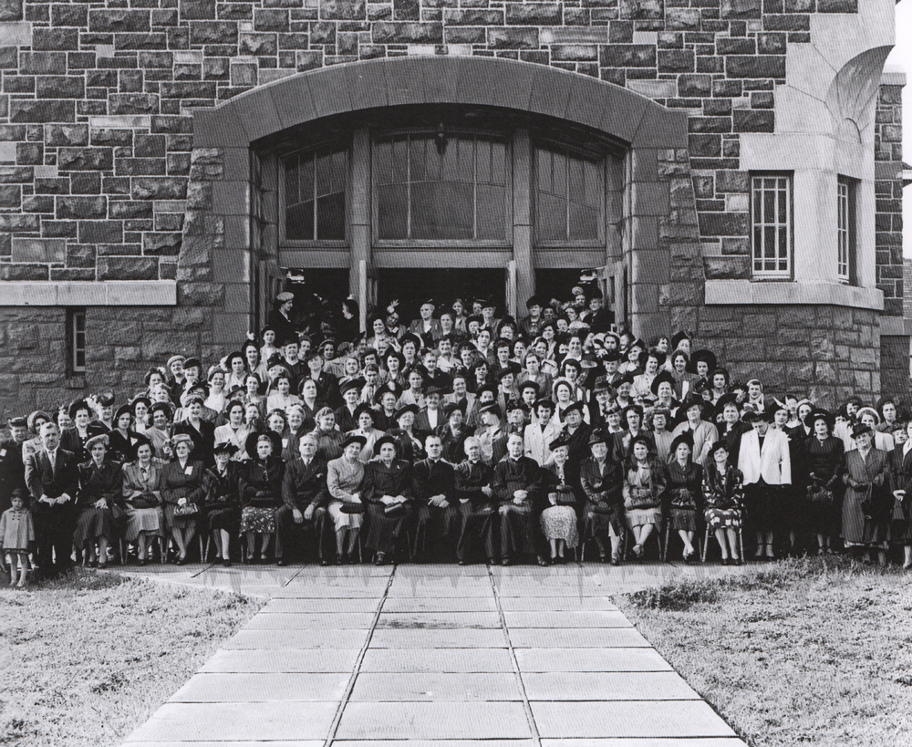 Old black and white photograph, long shot of a crowd of women surrounding a group of ecclesiastic dignitaries in front of a large stone church.