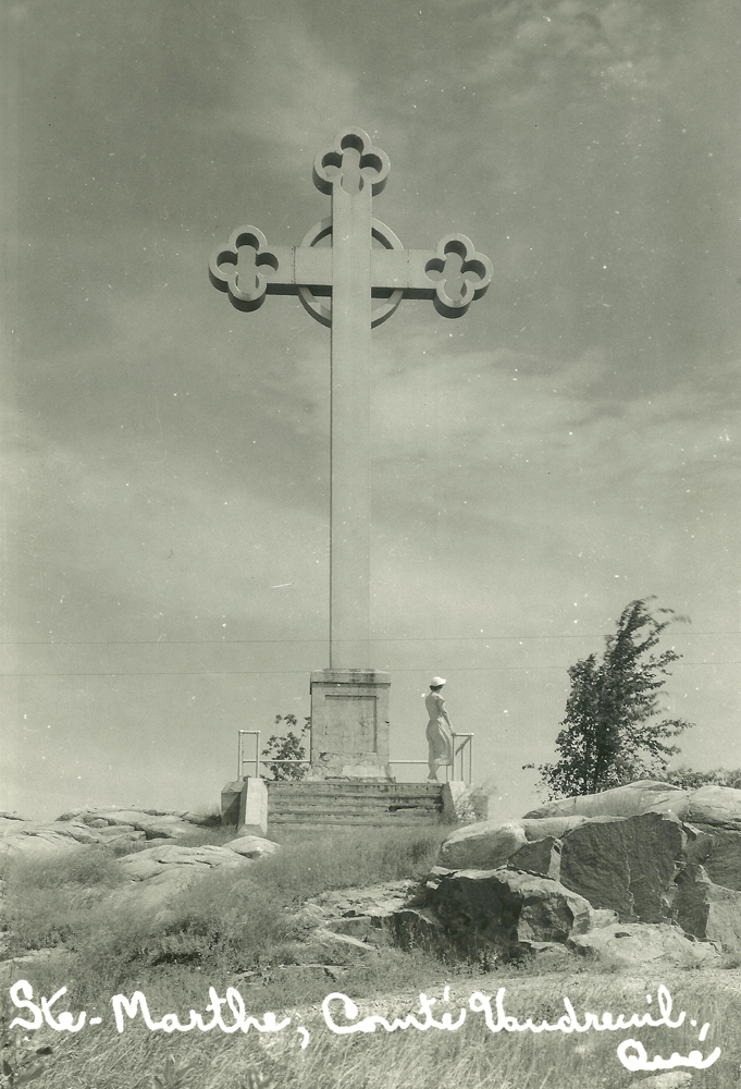 Black and white photograph, long shot of a very windy day, a woman looks out to the horizon as she stands under a huge stone cross built on a rocky promontory.