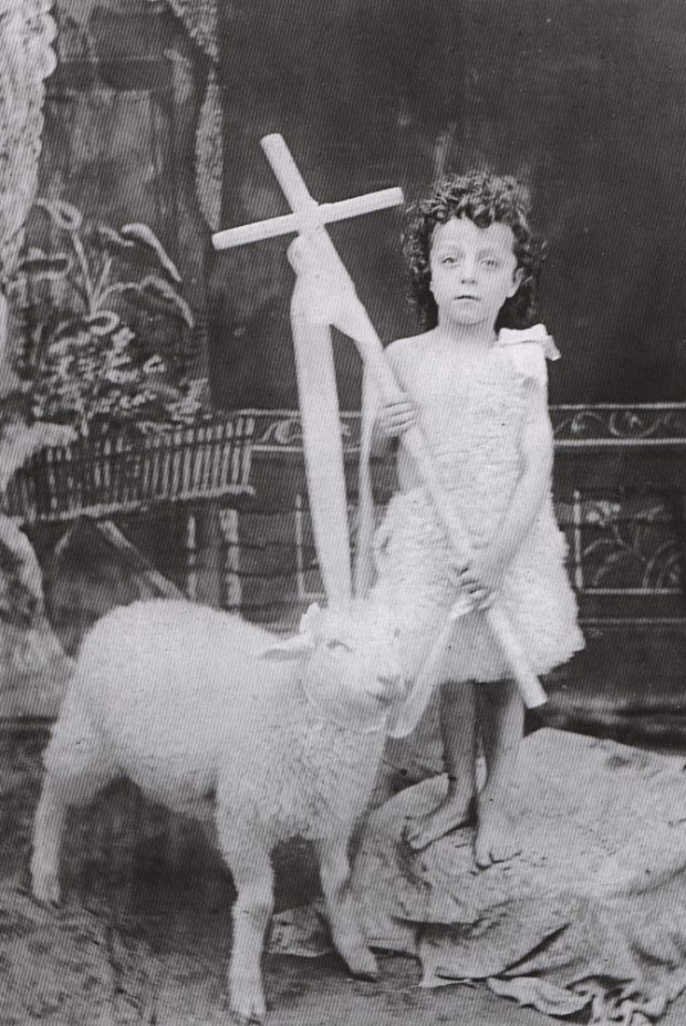 Old black and white photograph, close-up of a young boy holding a cross and wearing sheepskin, standing next to a small sheep.
