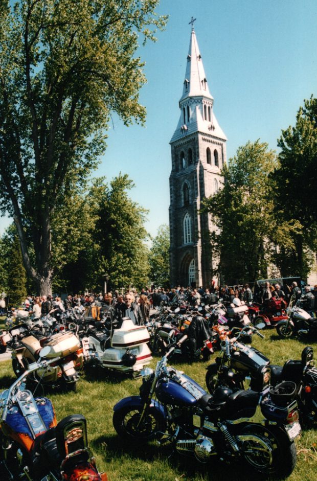 Color photograph, long shot of motorcycles and a crowd on a large lot surrounded by trees in front of a church.