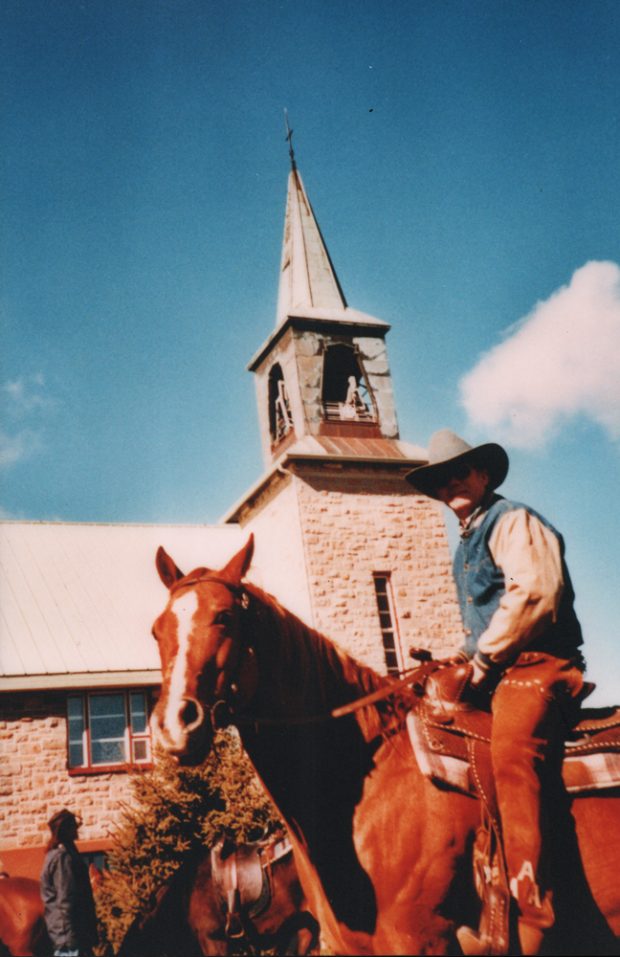 Color photograph, close-up of a cowboy on his horse next to a stone church and its steeple.