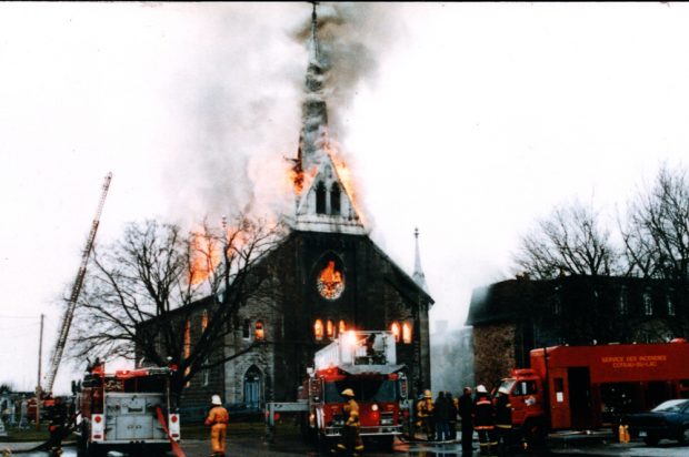 Color photograph, long shot of firefighters with a crane and many trucks, attempting to extinguish a church on fire whose steeple, roof and windows are in flames.