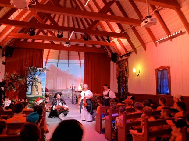 Color photograph, inside a small chapel transformed into a theater, children are sitting on wood pews and are listening to a theatrical representation with musicians in period costumes.