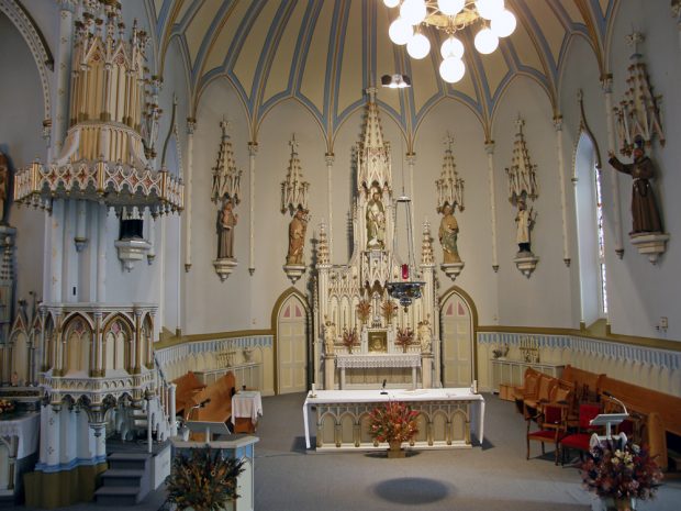 Interior view of a semicircle section of a church, long shot of walls with many religious plaster statues placed on wooden pedestals, above them are sculpted wooden canopies, in the center, liturgical furnishings.
