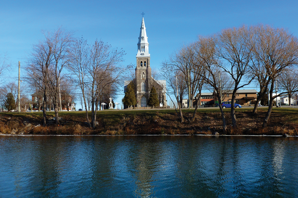 Color photograph, long shot, in the foreground, a river, riverside and trees, in the background, the façade of a large stone church and steeple.