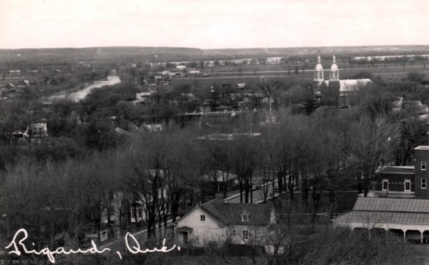 Old black and white photograph, wide aerial view of steeples breaking through trees and houses, in the background, a river.  